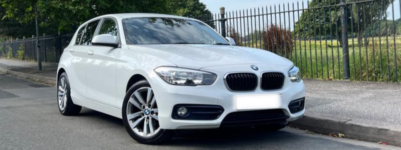 A white BMW 1 Series parked next to a park area in daylight.