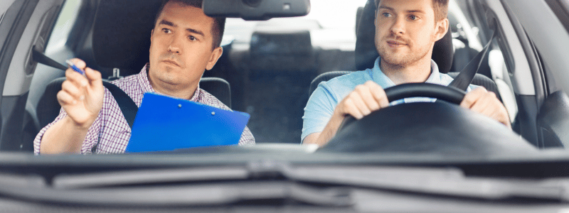 A driving instructor points in a direction whilst a driver watches on, representing driving lessons.