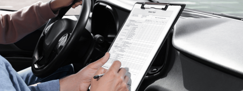 A person fills in a road test form whilst someone drives, symbolising a driving test.