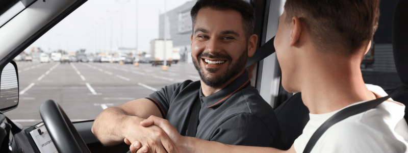 Smiling driving instructor shaking hands with a young driver inside a car, celebrating the completion of a driving test.