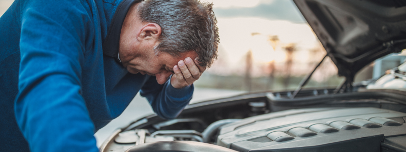 Man standing in front of a car with the hood open, looking frustrated with his hand on his forehead.