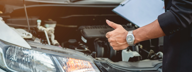 A close up of a car engine area, and someone stood in-front it with their thumb up whilst holding a checklist, representing a successful car check.