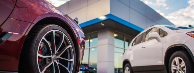 Close-up of cars parked outside a modern car dealership, highlighting vehicle wheels and dealership building with blue accents in the background.