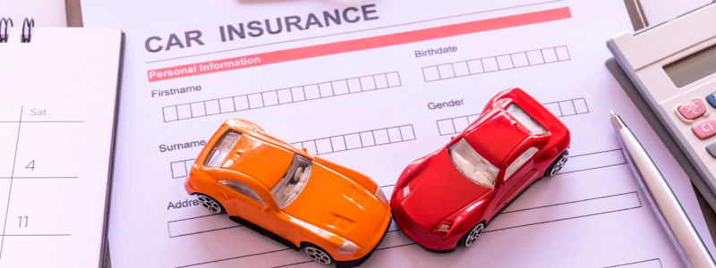 Two toy cars sat on top of a car insurance form, with a calculator and pen and a calendar in shot, symbolising setting up car insurance.