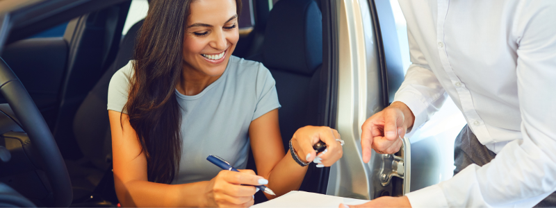 Someone signing a contract for a car whilst sat in the vehicle, and a business person outside the car pointing where to sign.