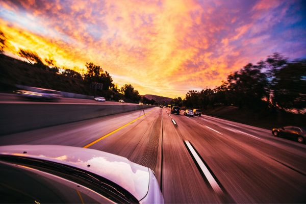 An overhead of the front of a car with a motorway ahead, and a sunset in the sky.