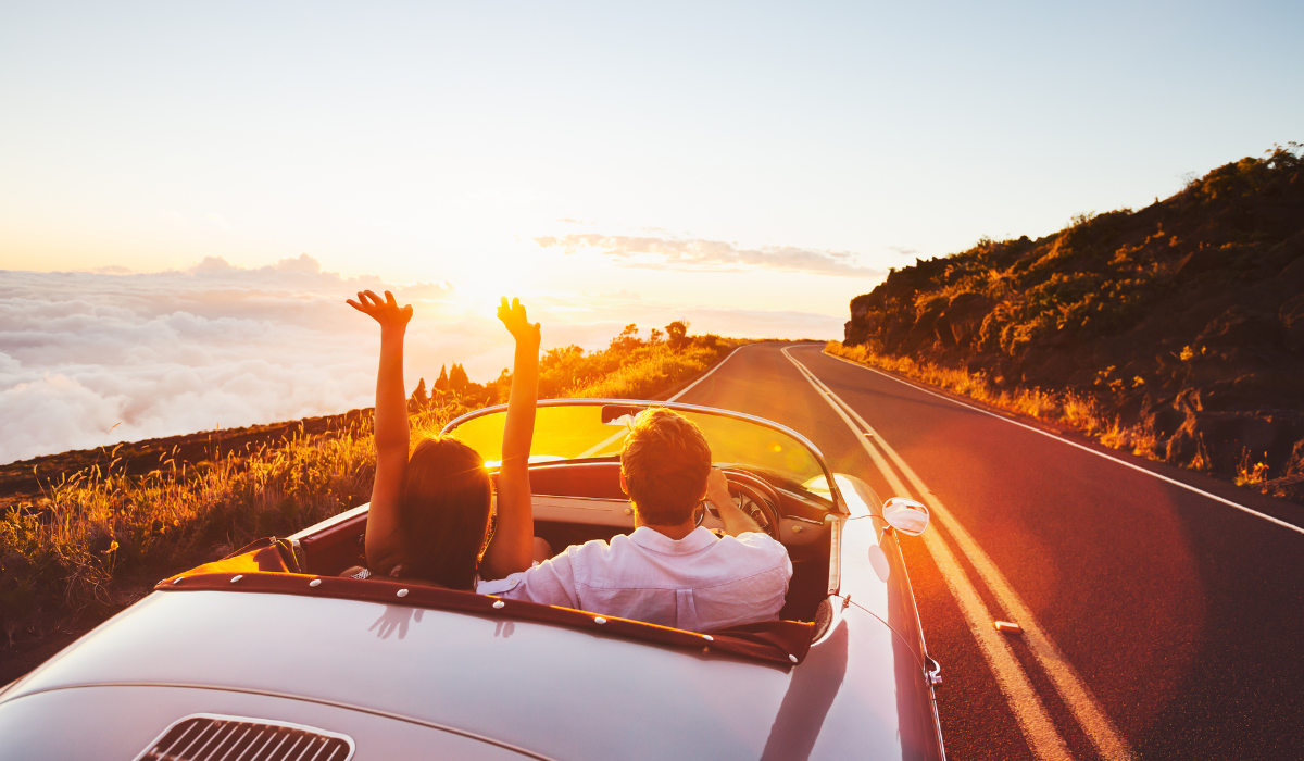 A couple driving their convertible car in the sun along a cliff-side road.