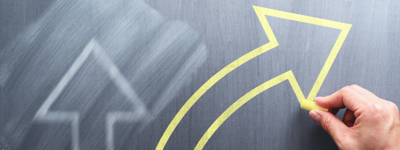 Hand drawing a bold yellow arrow on a chalkboard, symbolising growth, change, and upward direction, with a faint arrow outline in the background.