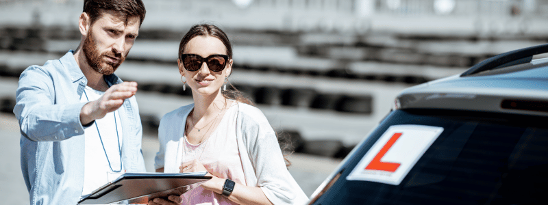 Driving instructor with a clipboard guiding a female learner driver next to a car with an 'L' plate, symbolising driving lessons.