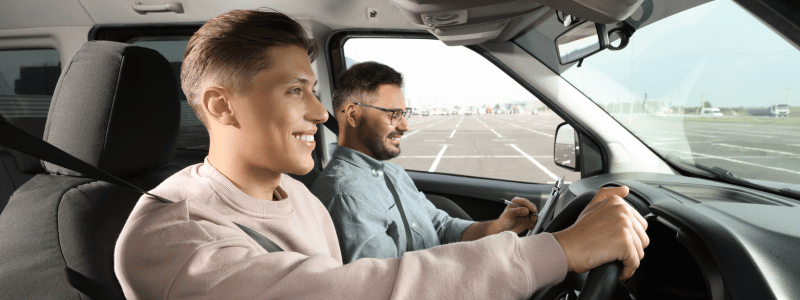 Young man smiling while driving with an instructor beside him during a driving test, symbolising confidence, learning, and road safety.