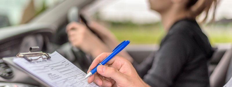 A passenger in a car making notes whilst someone else drives, likely in a driving test where an instructor is present.