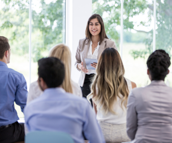 A female businessperson addressing a group of people in a work setting.