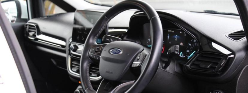 The interior of a Ford Fiesta with the driving seat in view. In shot is a steering wheel and dashboard with an infotainment system in place.