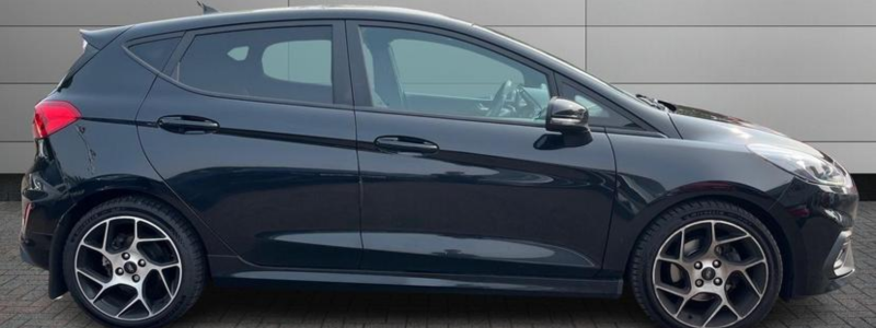 A side on view of a black Ford Fiesta ST, parked up against a grey wall.