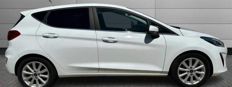 A white Ford Fiesta Titanium parked side-on against a plain grey wall, in an outside setting.