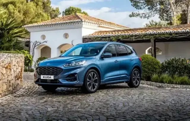 A blue Ford Kuga parked in front of a beautiful villa in a sunny, green area.