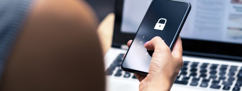 Person using a smartphone with a lock screen while working on a laptop, symbolising digital security, online privacy, and protection against fraud.