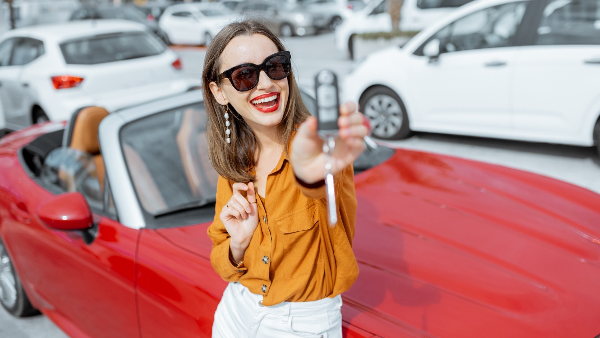 A happy woman at a car dealership showing her car keys to the camera with a red convertible in the background.