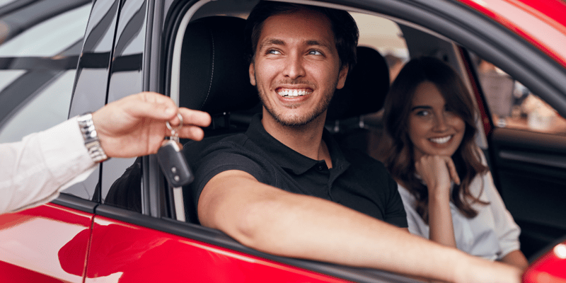 A person smiles whilst being handed car keys, with a woman sat in the passenger seat smiling and looking on.