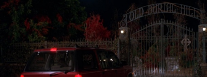 A red car parked in front of cemetery gates that read 'OLD BURIAL HILL'. A scene taken from the film Hocus Pocus.