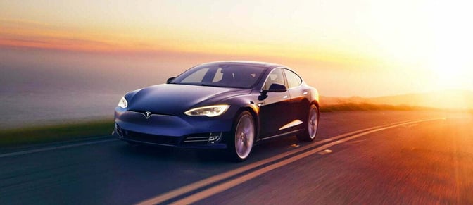 A blue Tesla Model S driving on a coastal road with a strong sunlight shining on the back of the car.