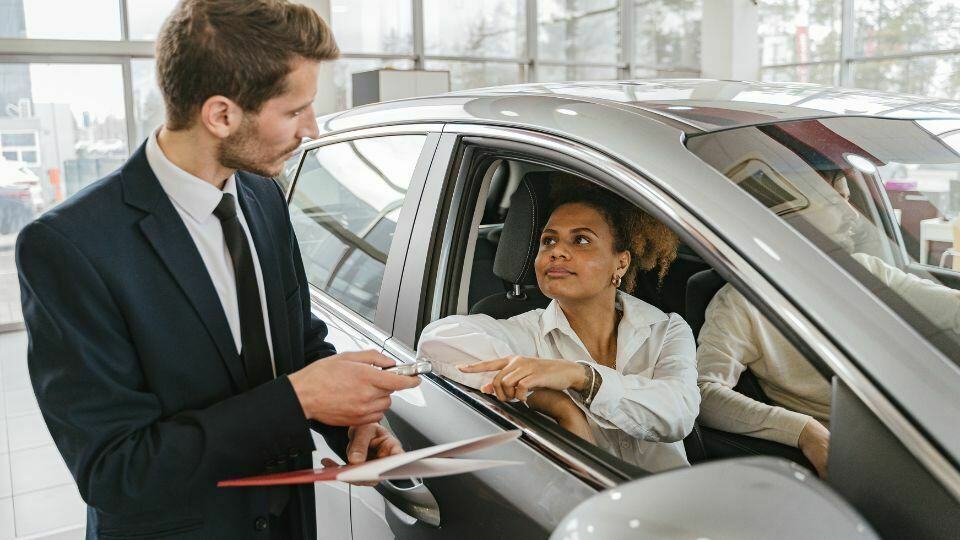 A woman sat in a car at a car dealership with a car salesman passing her the car keys