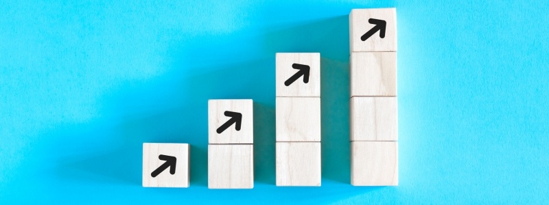 Wooden blocks stacked in ascending order with black arrows pointing up, against a blue background, representing growth, progress, and upward trends.