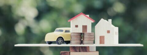 Two model houses, a toy car and two stacks of coins sit central on a wooden scale.