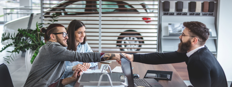 Smiling couple shaking hands with a salesman at a car dealership, symbolising a successful car purchase and positive customer service experience.