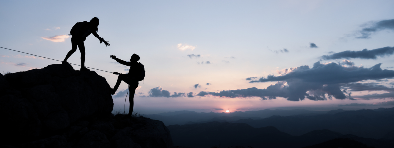 A climber reaches below to help someone up over a hill, representing teamwork and support.