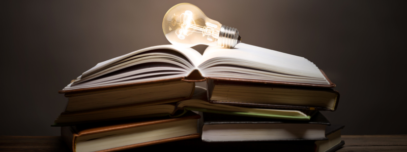 Stacks of open books with a lit light bulb on top, symbolising knowledge and thought.