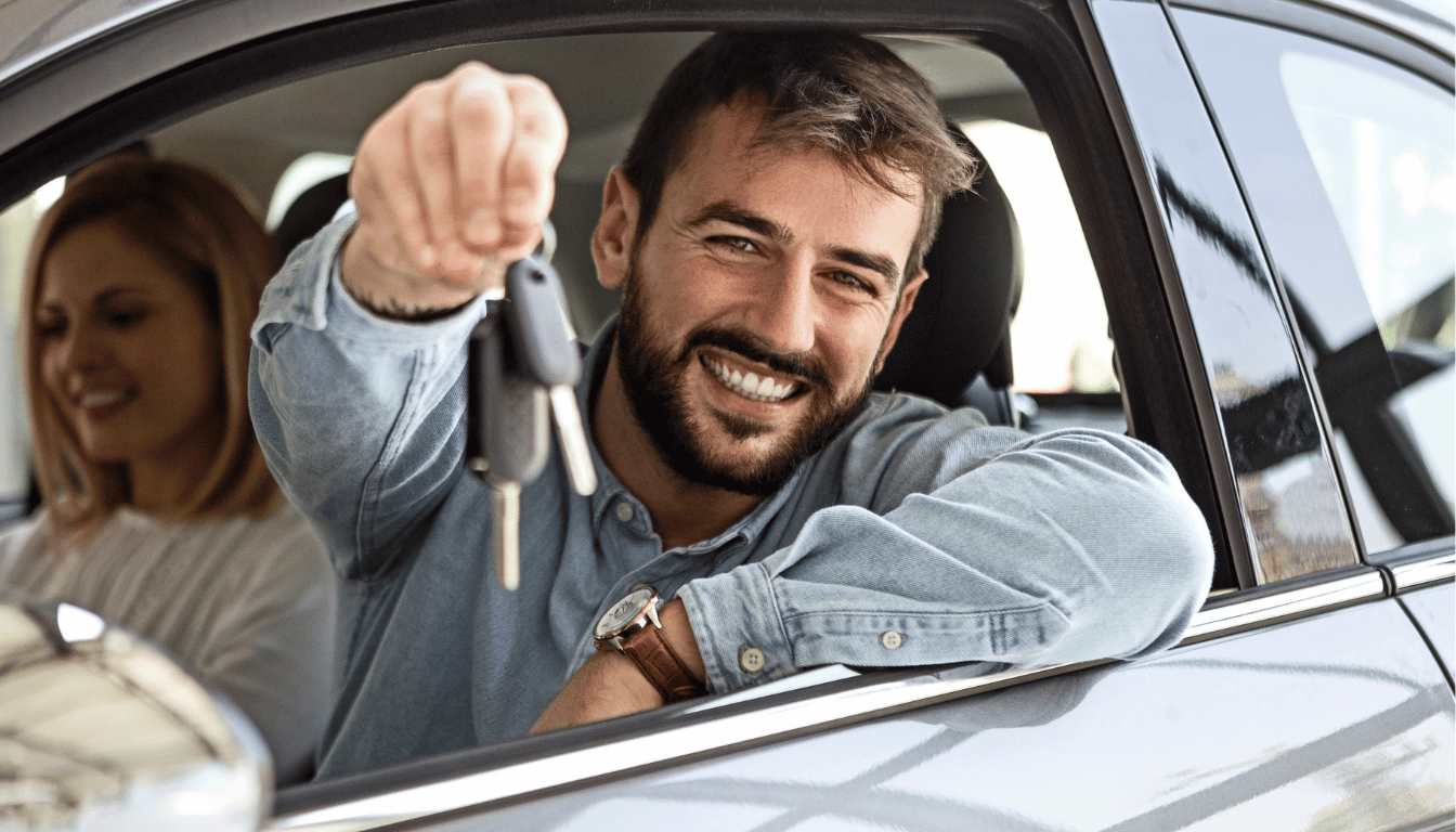 A person holds car keys and smiles whilst sat in their car.