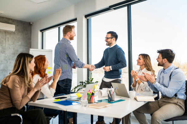 Two people shake hands stood up in a business setting whilst others sit around them and applaud.