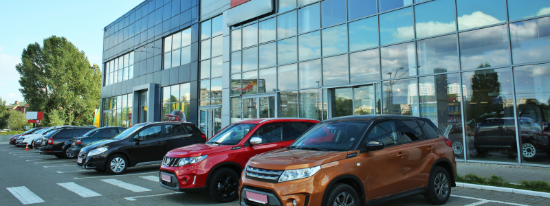 Line of parked cars outside a modern glass-walled dealership building, showcasing a variety of vehicles on display.