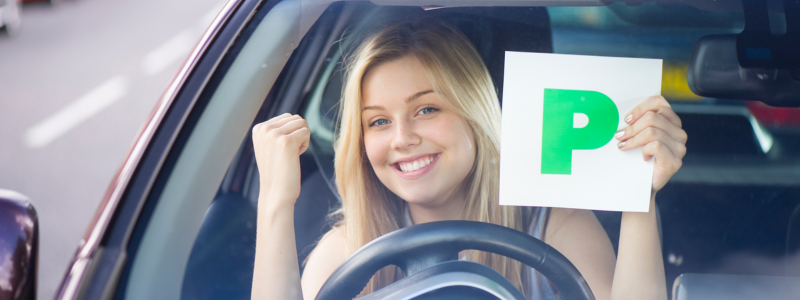 A person smiling and celebrating in their car whilst holding up a P sticker, indicating that they have just passed their test.