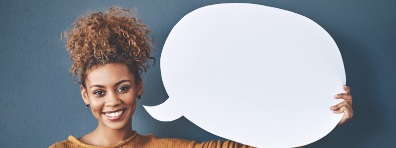 A person smiling at the camera whilst holding up a white speech bubble cut out, symbolising them speaking and offering their opinion.