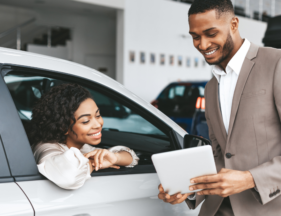 A car salesman shows a tablet device to a woman sat in a white car in a showroom setting.