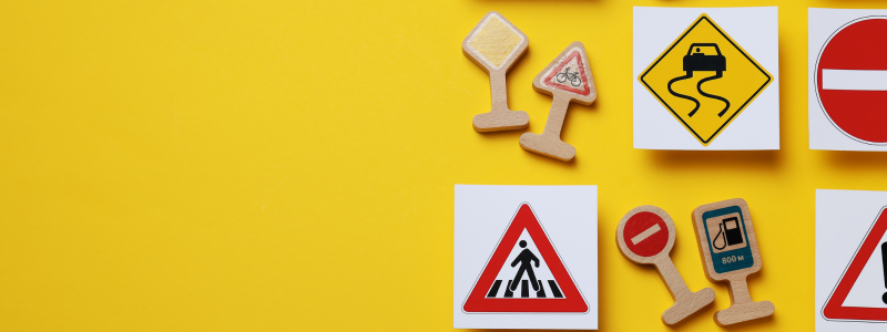 Road signs on white backgrounds and as cut outs, sat together in the right hand side of the picture, with a yellow background throughout.