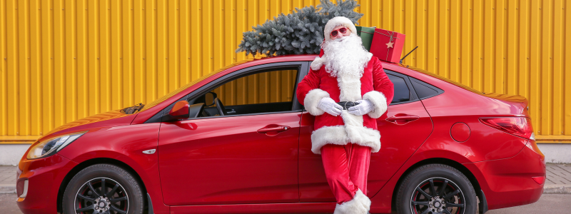 Santa in front of a red car with a Christmas tree and presents on top.