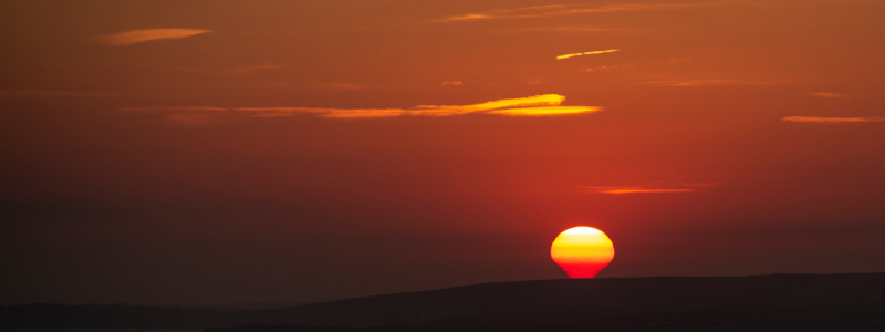 A dark red sky, with an orange and red sun disappearing below a hillside, symbolising sunset and the end.