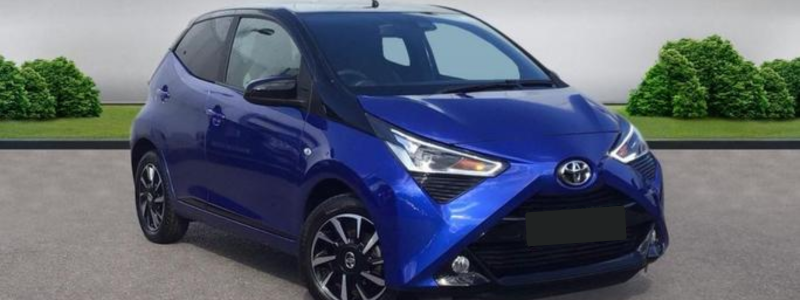 A blue Toyota Aygo parked in front of a grey wall with small greenery in front of it.