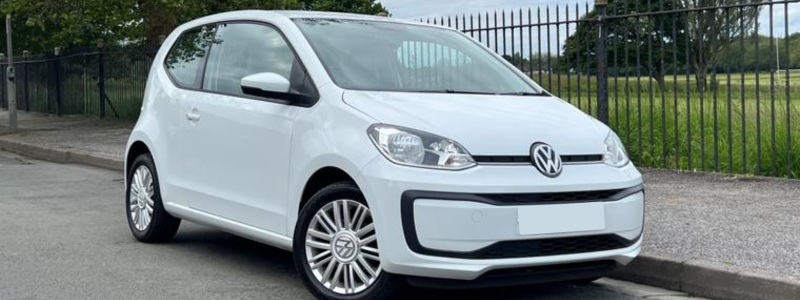 A white Volkswagen Up parked by the side of a road outside of a park, in a day time setting.