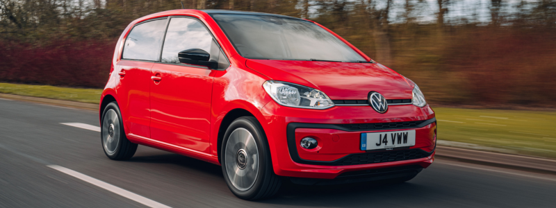 A red VW Up driving on a road with greenery in the background.