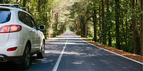 A white SUV driving on a straight road with trees either side and sun shining down.