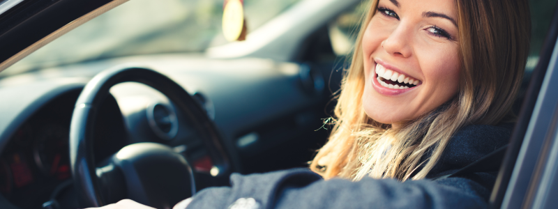 A smiling woman looking at the camera whilst sat in a driving seat. 