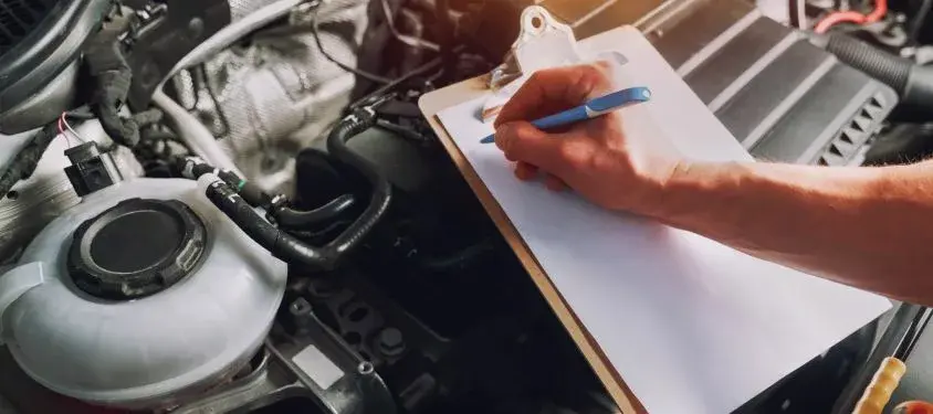 A person writes on a clipboard that's leaning on the engine of the car.